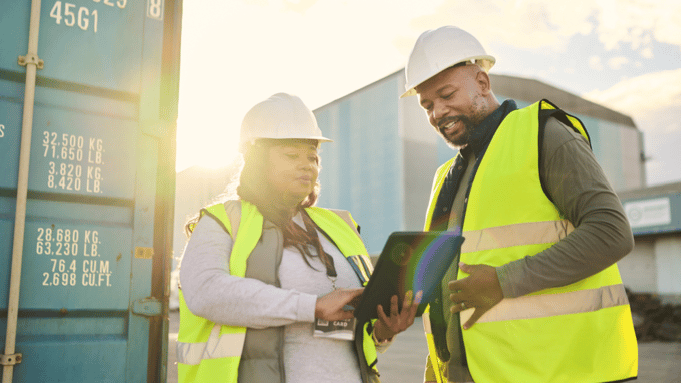 two male and female african workers looking at a tablet with shipping containers in the background