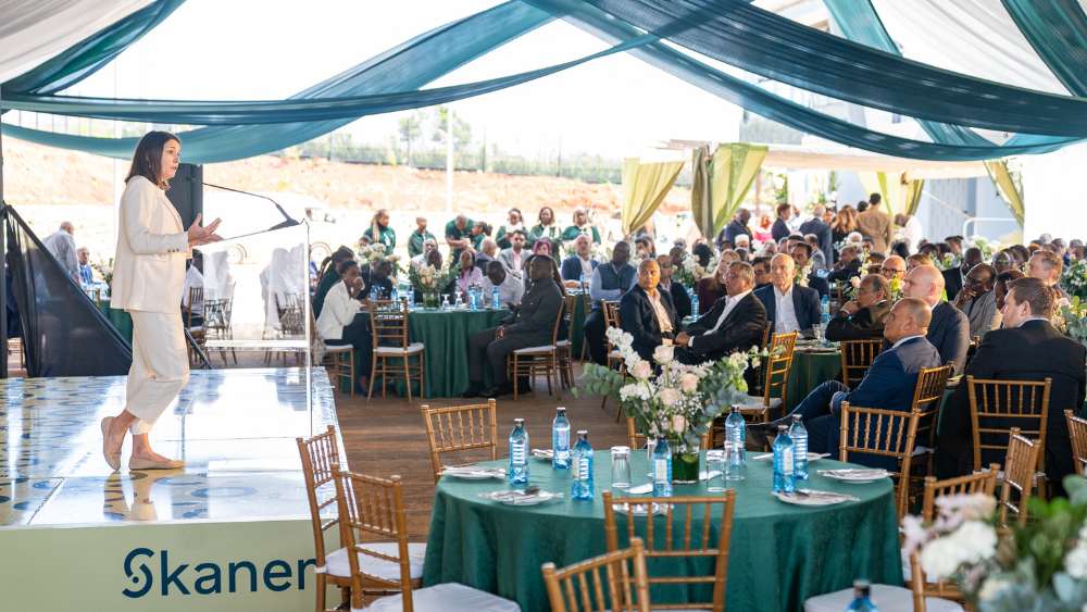 A woman in a white suit addressing an audience at a formal event, with tables set under a tent decorated with green and white drapes.