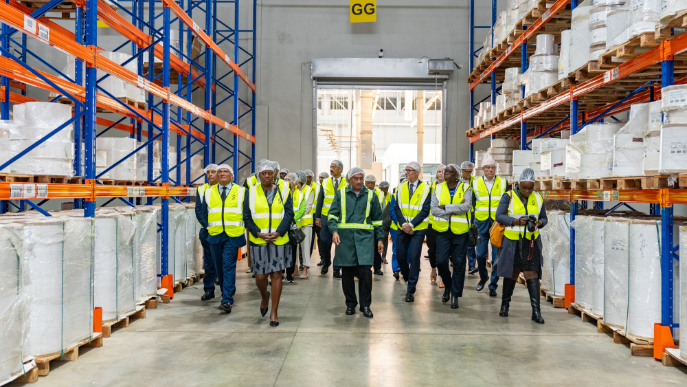 A group of visitors in reflective vests and safety gear touring a modern warehouse facility filled with stacked pallets and shelves.